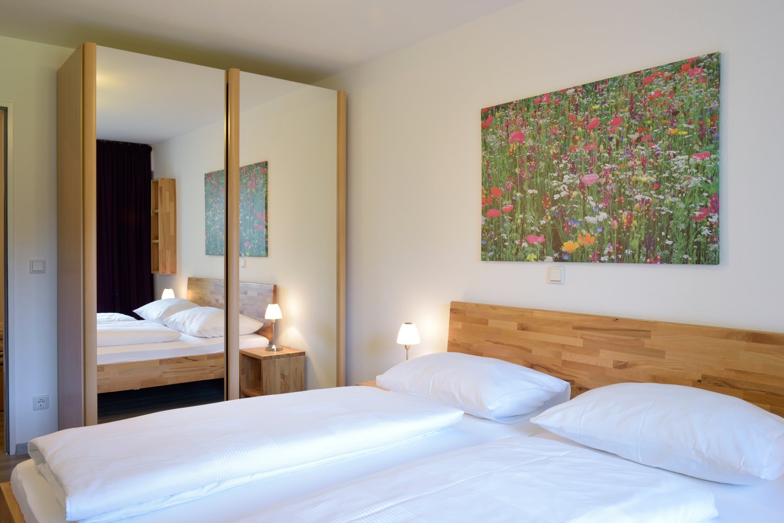 Modernes Schlafzimmer in der Ferienwohnung des Die Gams Hotel Resort in Bad Hindelang mit Doppelbett, Spiegelkleiderschrank und dekorativem Blumenbild.