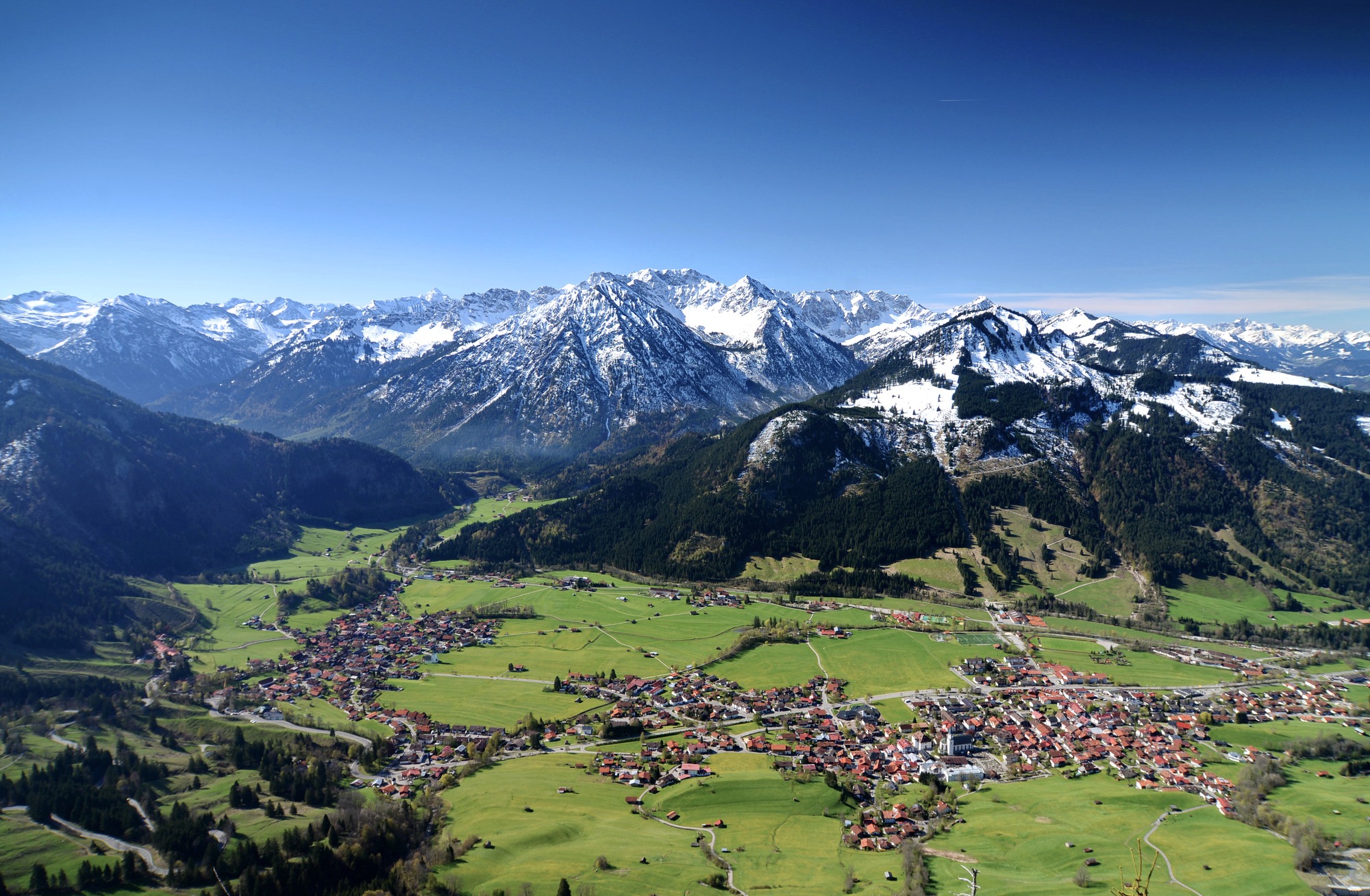 Panoramablick auf Bad Hindelang im Allgäu mit malerischem Tal, verschneiten Alpen und tiefblauem Himmel.