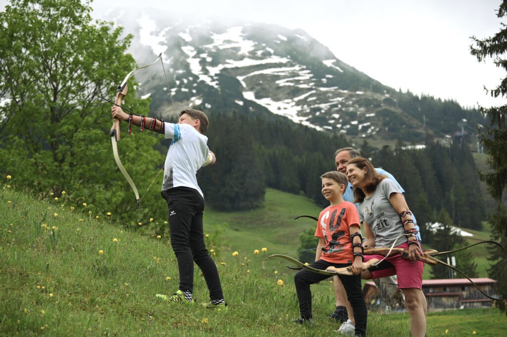 Familie beim Bogenschießen in Bad Hindelang im Allgäu vor alpiner Bergkulisse mit Wiesenhang und schneebedecktem Gipfel im Hintergrund.