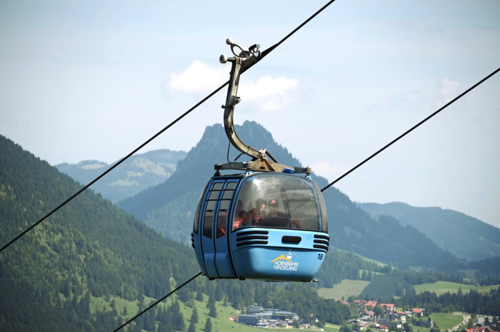 Blau gondelnde Hornbahn Hindelang mit Ausblick auf das grüne Tal und die Allgäuer Alpen bei sommerlichem Wetter.