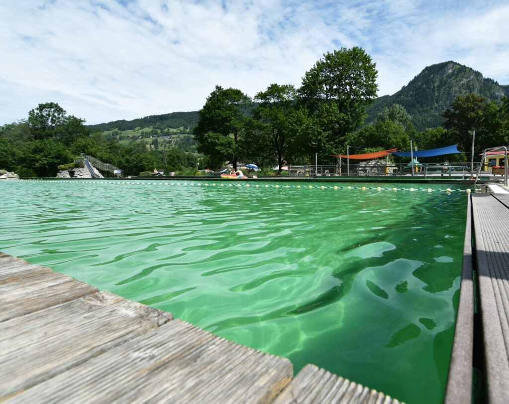 Naturfreibad in Bad Hindelang im Allgäu mit grünem Wasser, Holzstegen, Spielbereich und Blick auf die umliegende Berglandschaft.