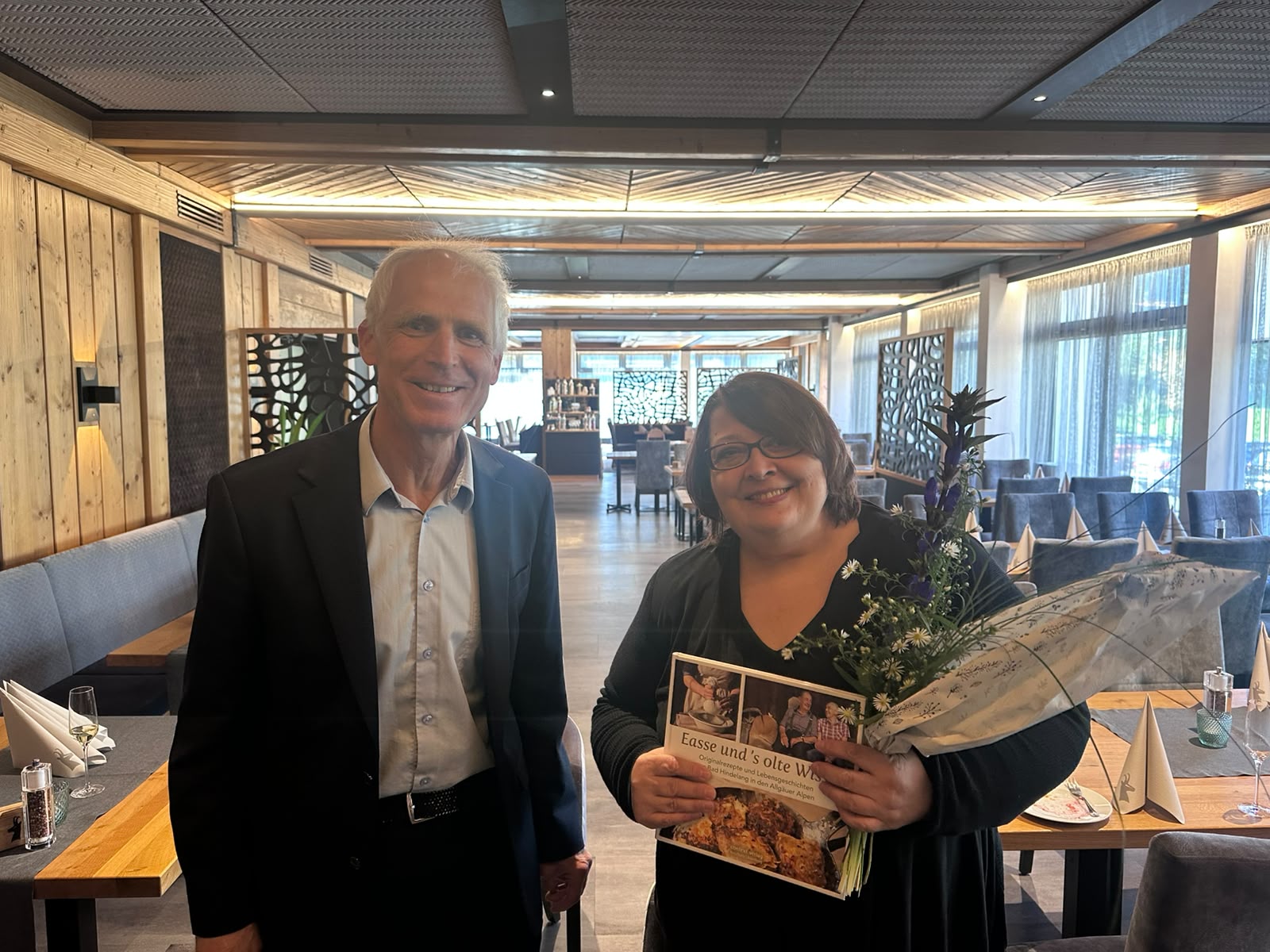 Elisabeth Bichler mit Blumen und Buch neben Max Hillmeier, Leiter von Bad Hindelang Tourismus, im Restaurant der GAMS
