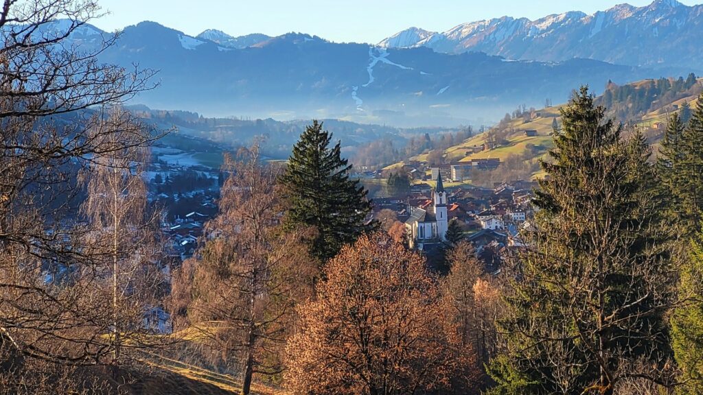 Herbstlich gefärbte Bäume am Hang mit Blick auf Bad Hindelang, Kirchturm und die Allgäuer Berge