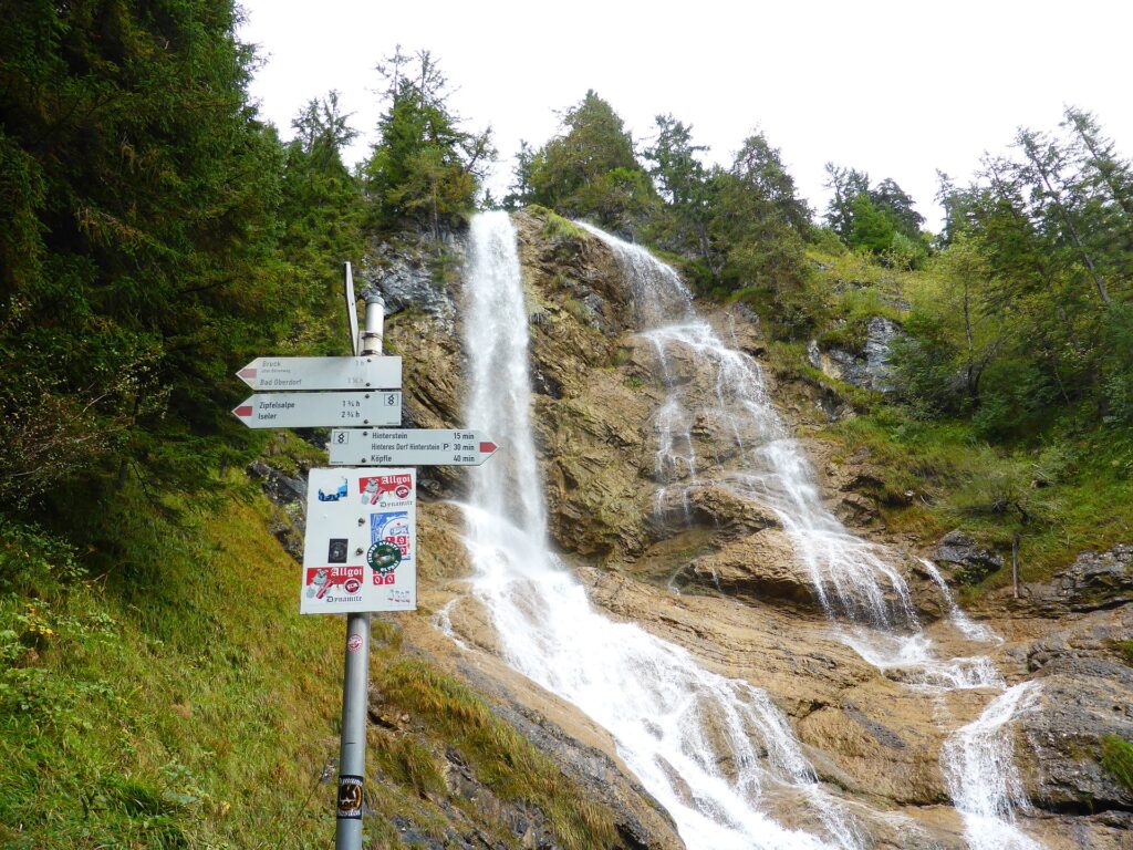 Zipfelsbachfälle bei Hinterstein: Wasserfall über Felsstufen, davor ein Wegweiser am Wanderweg