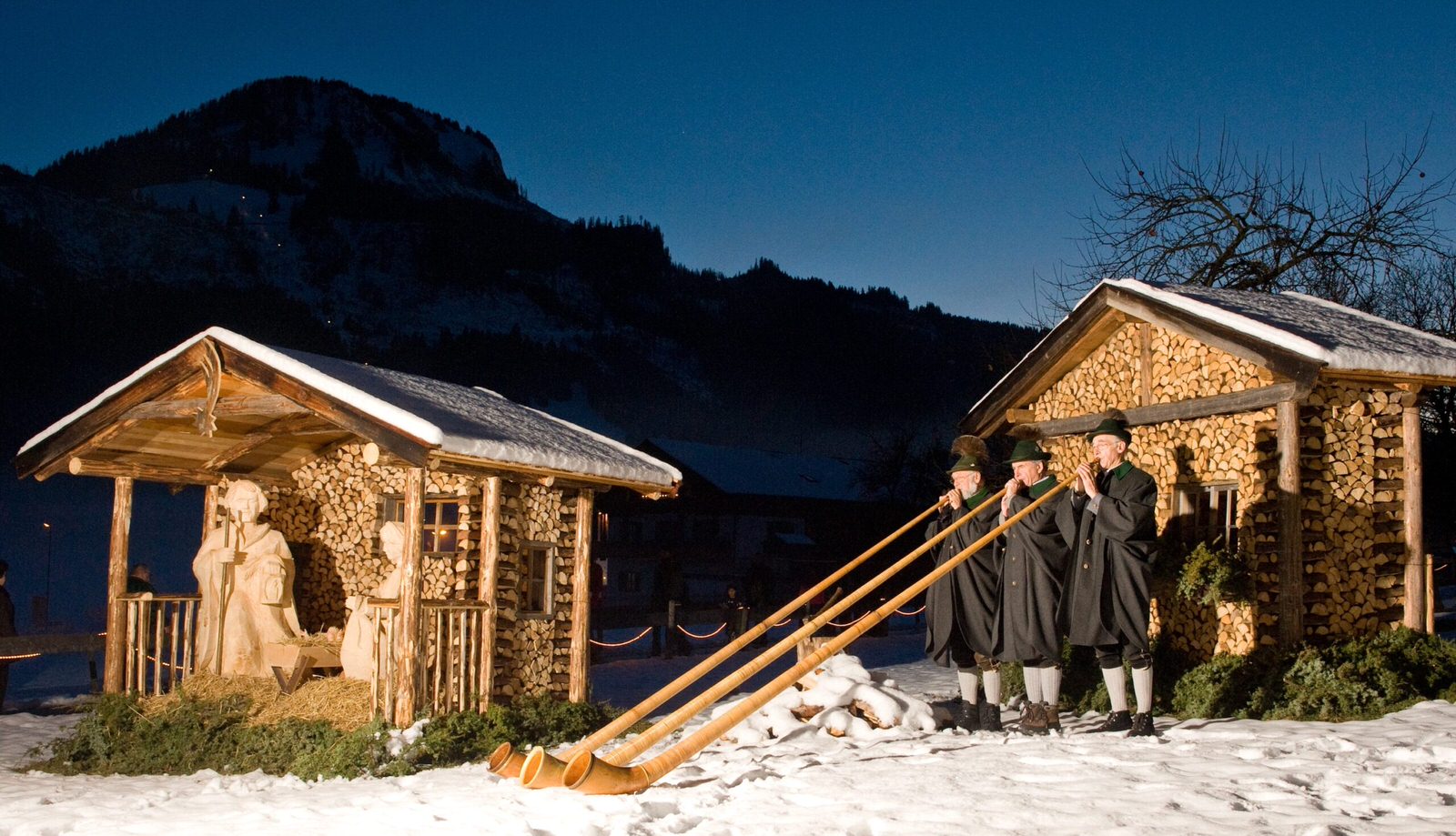 Holzkrippe im Schnee, daneben Alphornbläser vor den Bergen in der Abenddämmerung