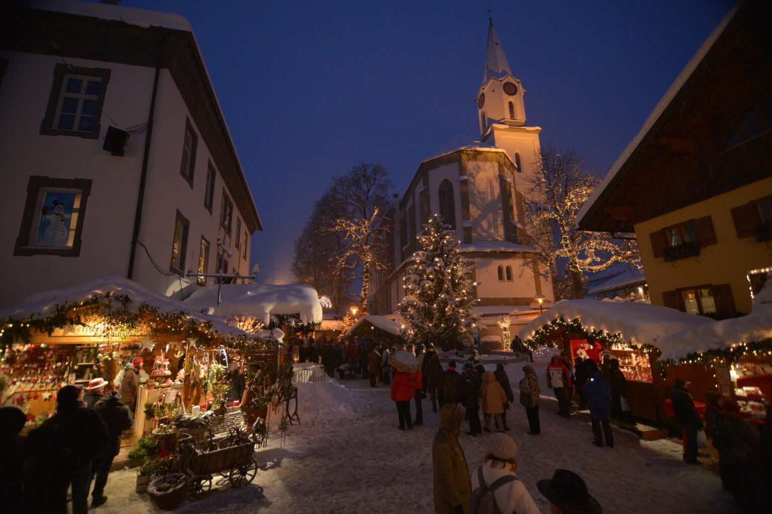 Erlebnis-Weihnachtsmarkt Bad Hindelang am Abend: beleuchtete Hütten im Schnee, dahinter die Pfarrkirche