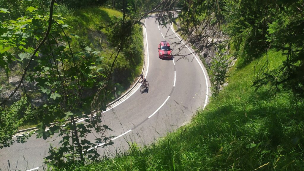 Blick durch Laub auf eine Kehre der Jochstraße mit einem Rennradfahrer und einem Auto