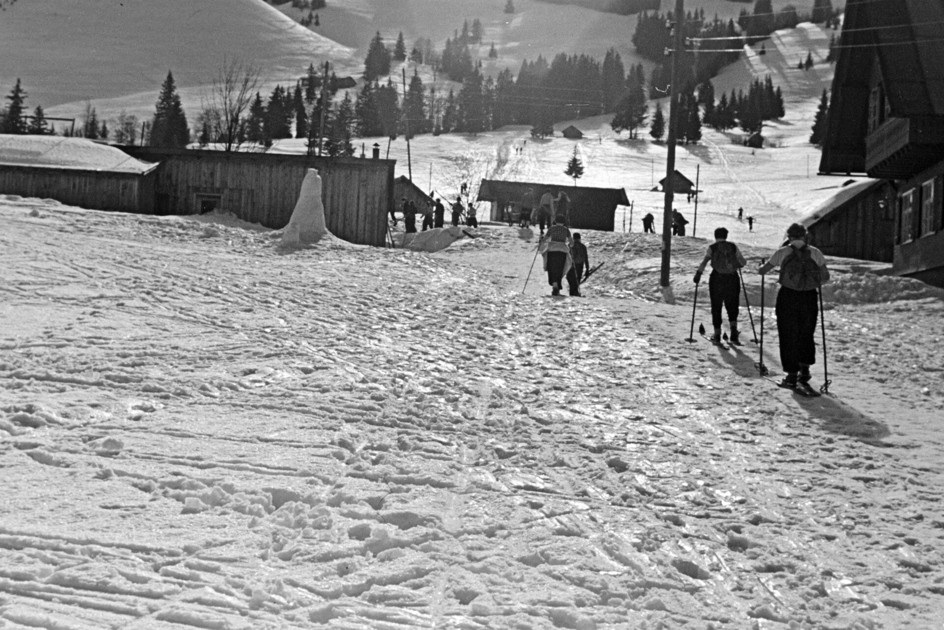 Historisches Schwarzweißfoto: Skifahrer steigen in Oberjoch zwischen Holzhäusern einen verschneiten Hang hinauf