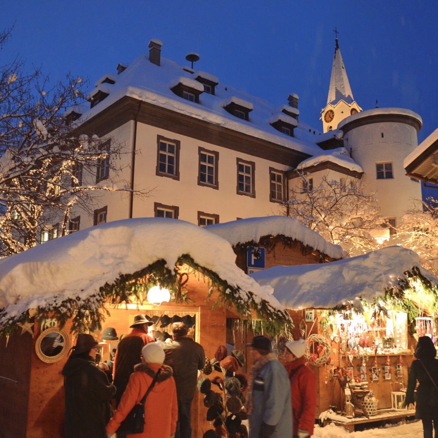 Stimmungsvoller Weihnachtsmarkt in Bad Hindelang im Allgäu mit festlich geschmückten Holzhütten, Lichterketten und verschneiter Kulisse am Abend.